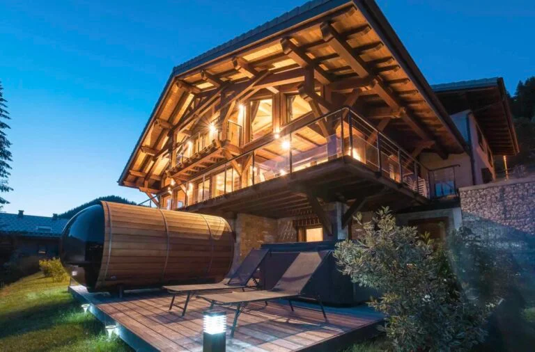 Façade éclairée d’un Chalet Bovard avec sauna extérieur à la tombée du jour